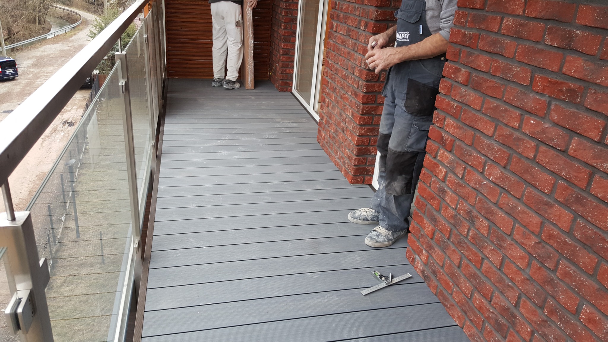 Montaż szarej deski kompozytowej na balkonie z balustradą ze szkła, widoczny pracownik w ubraniu roboczym i poziomica.