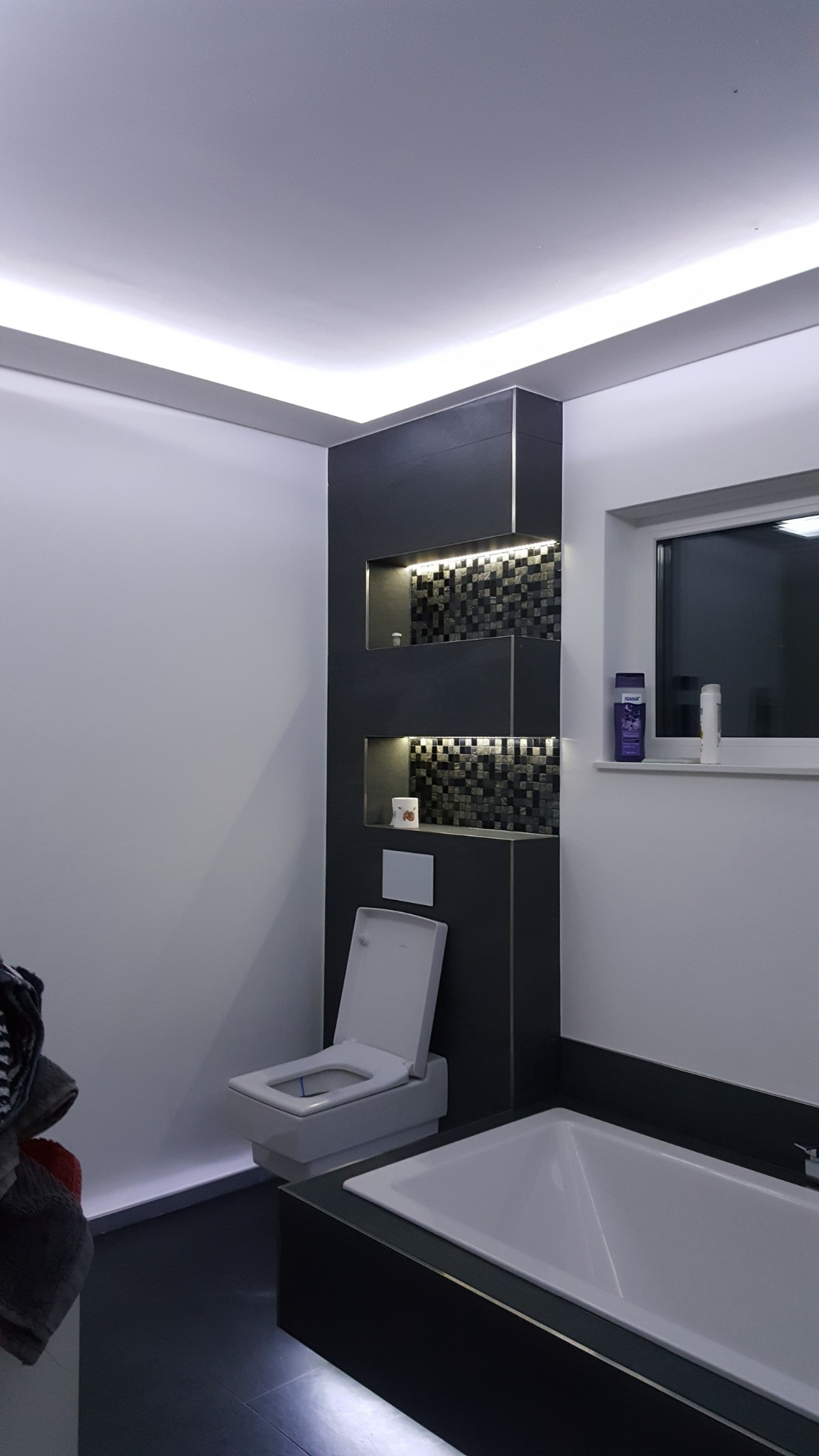 Nowoczesna łazienka z podświetlanymi wnękami wyłożonymi mozaiką, białą wanną i toaletą, oświetleniem LED podsufitowym.