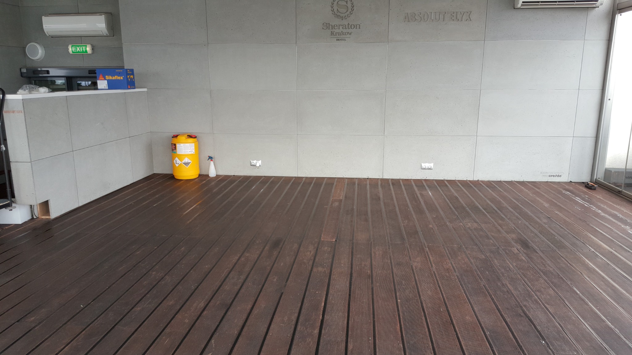 Drewniana podłoga tarasu zewnętrznego przed betonową ścianą z logo Sheraton Krakow i Absolut Elyx, żółty kanister i butelka ze spryskiwaczem w rogu.