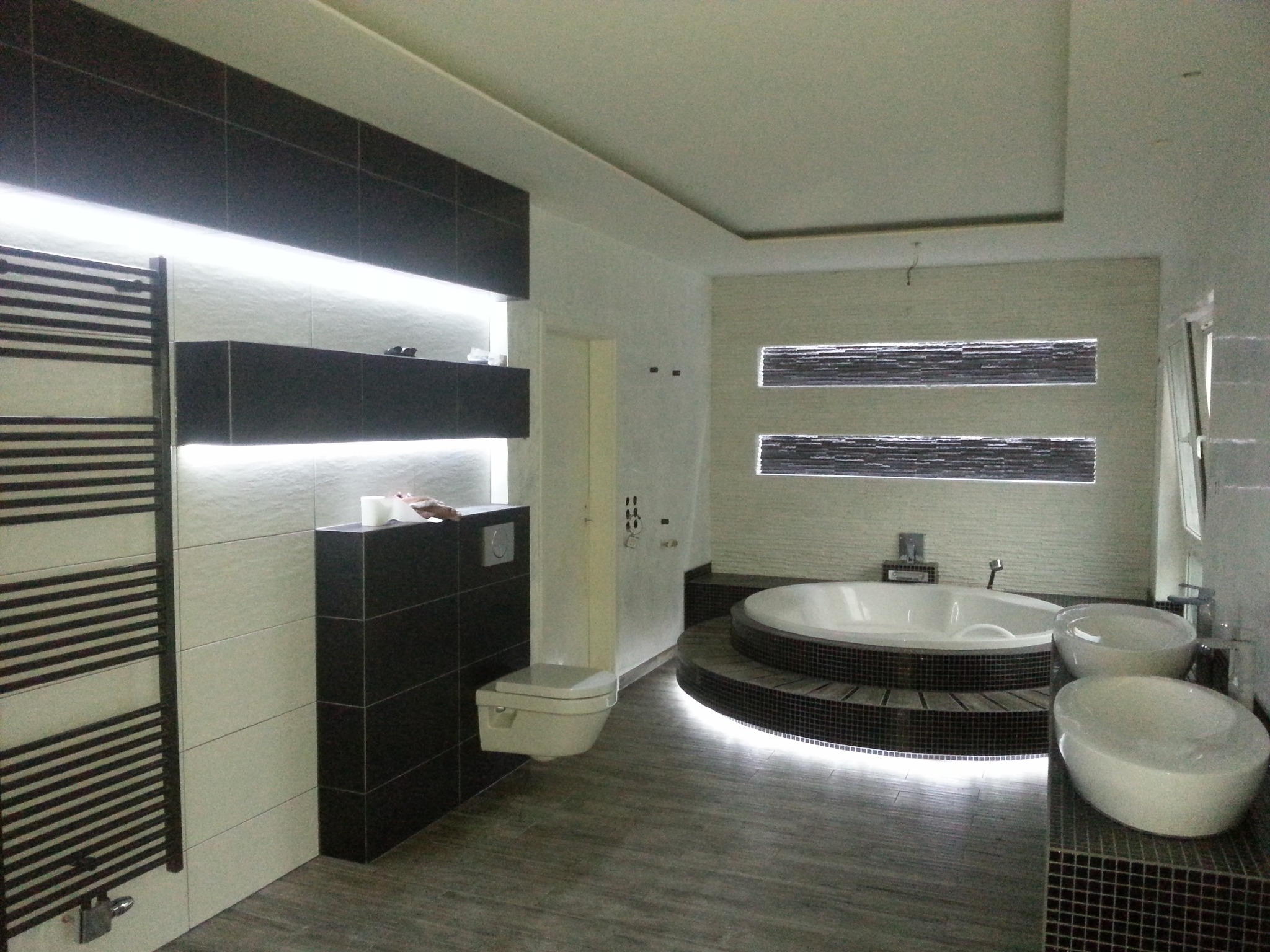 Nowoczesna łazienka z okrągłą wanną na podeście z mozaiki, podświetlana listwą LED, grzejnikiem drabinkowym i podwieszaną toaletą w kolorach bieli i ciemnego brązu.