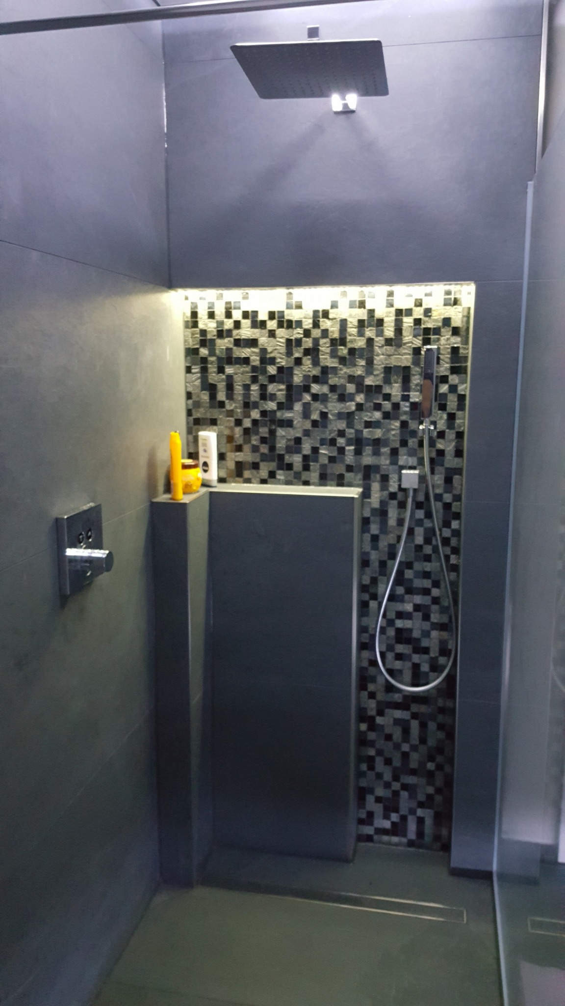 Nowoczesna kabina prysznicowa z szarymi płytkami, mozaiką w strefie prysznica i deszczownicą, oświetleniem LED oraz widocznymi kosmetykami na półce.