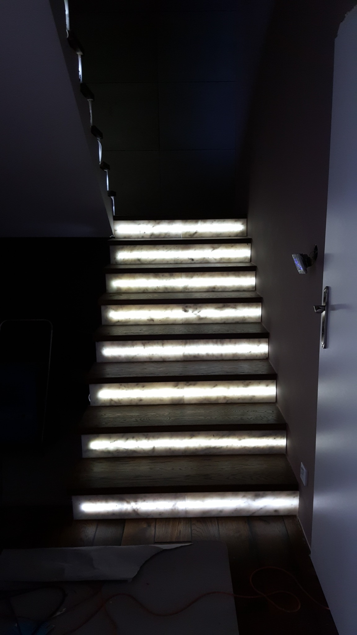 Schody z podświetleniem LED, ciemne drewniane stopnie i marmurowe podstopnice emitujące jasne światło, widok z dołu.