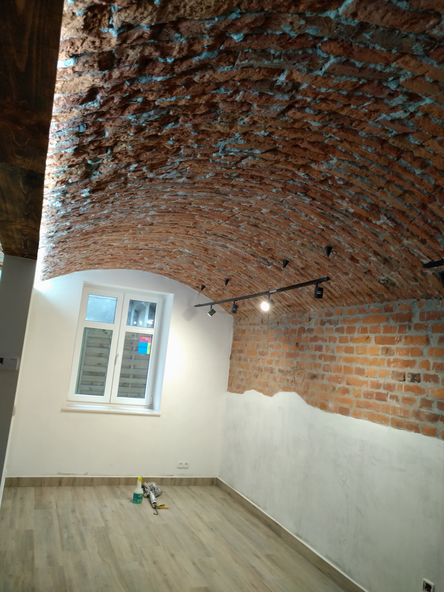 Pokój z odrestaurowanym sklepieniem z cegły, częściowo otynkowanymi ścianami i drewnianą podłogą; widoczne narzędzia i butelka płynu do czyszczenia.