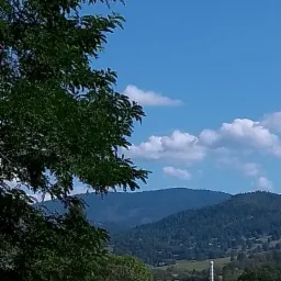 Pejzaż górski z bujną zielenią drzew w lewym rogu, błękitne niebo z obłokami w tle i widocznym kominem w oddali.