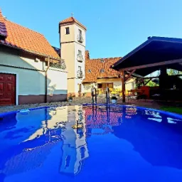 Niebieski basen ogrodowy z odbiciem wieżyczki budynku w tle i zadaszoną strefą relaksu z grillem.