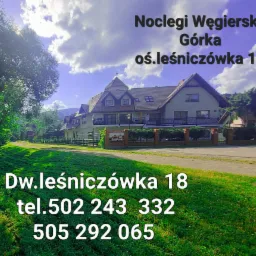 Dwór Leśniczówka 18 w Węgierskiej Górce, widok z zewnątrz na budynek z ogrodem i tablicą informacyjną, w tle góry i niebo z chmurami.