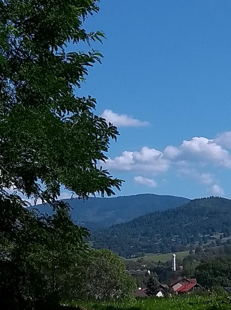 Pejzaż górski z bujną zielenią drzew w lewym rogu, błękitne niebo z obłokami w tle i widocznym kominem w oddali.