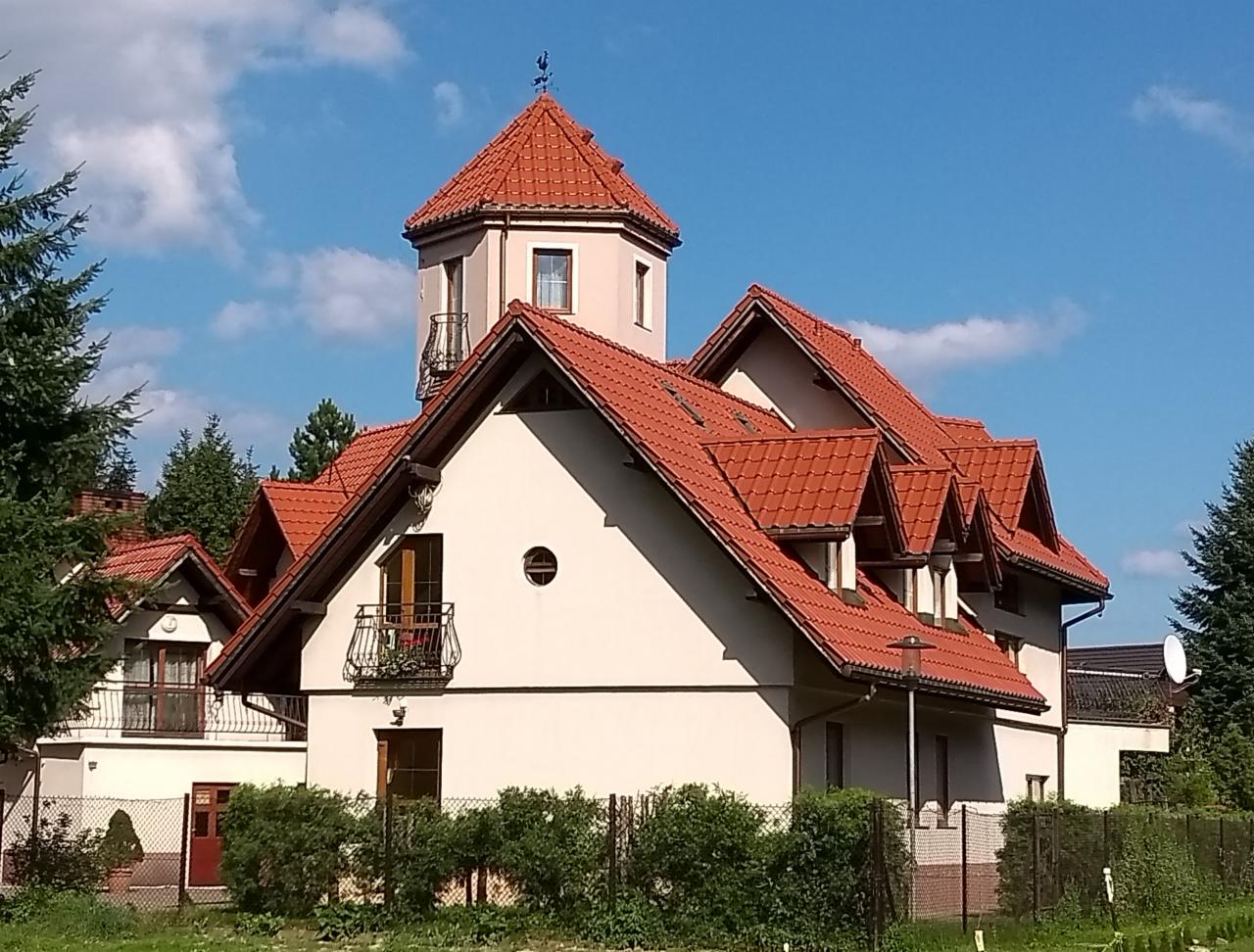 Elegancki dom z wieżyczką i balkonem, pokryty czerwoną dachówką, otoczony zielenią. Ujęcie w słoneczny dzień, z widocznym fragmentem ogrodzenia.