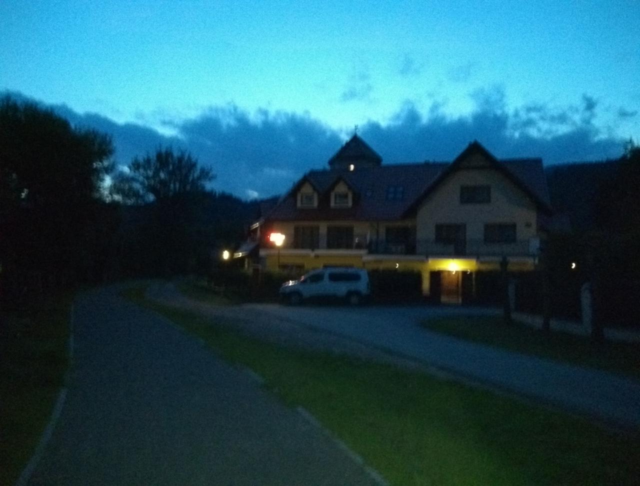 Dwupiętrowy budynek z poddaszem i balkonem oświetlony wieczorem, z zaparkowanym samochodem na podjeździe, otoczony zielenią i drzewami.