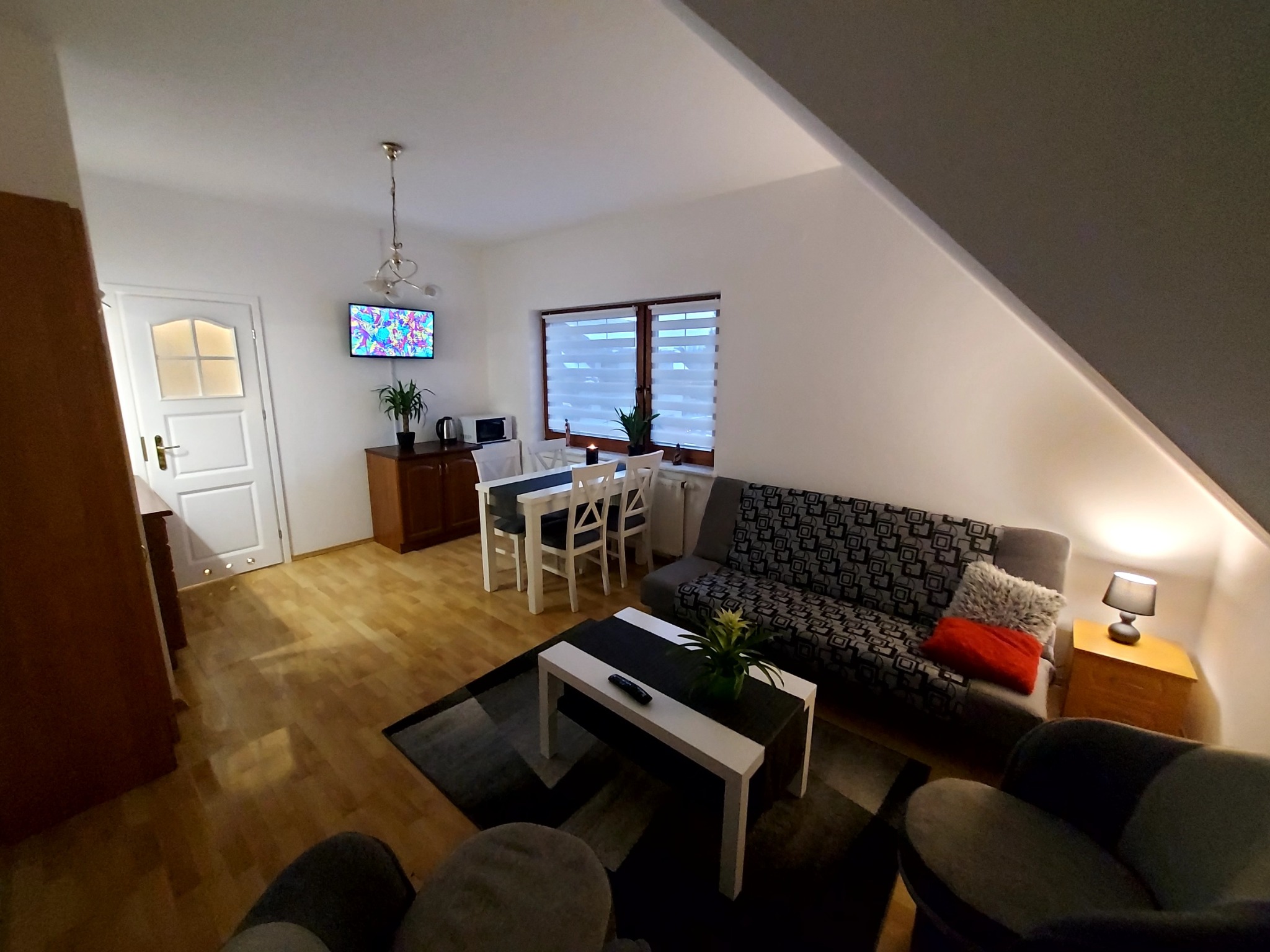 Jasne wnętrze pokoju z drewnianą podłogą, skośnym sufitem, stołem jadalnym, sofą z wzorem geometrycznym, stolikiem kawowym i telewizorem z abstrakcyjnym obrazem.