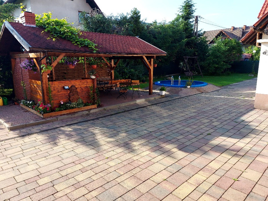 Drewniana altana z czerwonym dachem, porośnięta zielenią, z kwiatami w donicach, obok mały basen ogrodowy i plac wyłożony kostką brukową.