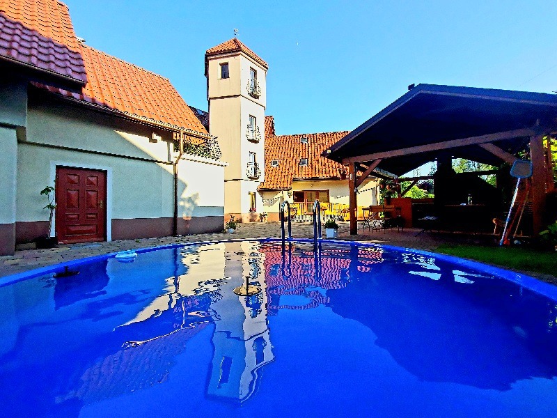 Niebieski basen ogrodowy z odbiciem wieżyczki budynku w tle i zadaszoną strefą relaksu z grillem.