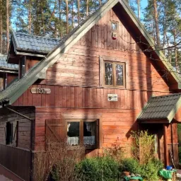 Dwupiętrowy, brązowy dom z drewna z szarym dachem w leśnej scenerii, z widocznym numerem 180A i tabliczką firmy ochroniarskiej.