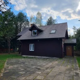 Ciemnobrązowy dom z brązowym dachem z dachówki, widok z zewnątrz na tle drzew i pochmurnego nieba, z betonowym podjazdem z kostki.