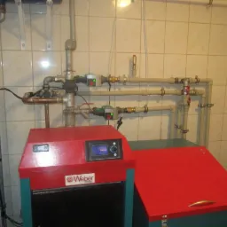 Czerwono-zielony piec Weber z podłączonymi rurami instalacji grzewczej na tle jasnych, kwadratowych płytek w kotłowni.