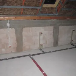 Surowy stan poddasza z instalacją hydrauliczną w ścianach i na podłodze, widoczne rury i otwory, przygotowane do dalszych prac wykończeniowych.