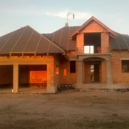 Surowy stan domu jednorodzinnego z częściowo położonym pokryciem dachowym. Widoczne elementy konstrukcji i pustaki ceramiczne. Dom w trakcie budowy.