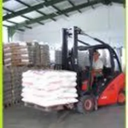 Wózek widłowy transportuje paletę z workami w magazynie. Widoczny operator wózka i rzędy ułożonych worków w tle.