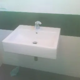 Biała, prostokątna umywalka zamontowana na ścianie z chromowaną baterią, widoczna na tle białych i zielonych płytek, z niebieską szmatką leżącą na podłodze.