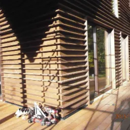 Fragment elewacji budynku z drewnianymi żaluzjami, widoczne narzędzia i buty robocze na drewnianym tarasie w słoneczny dzień.