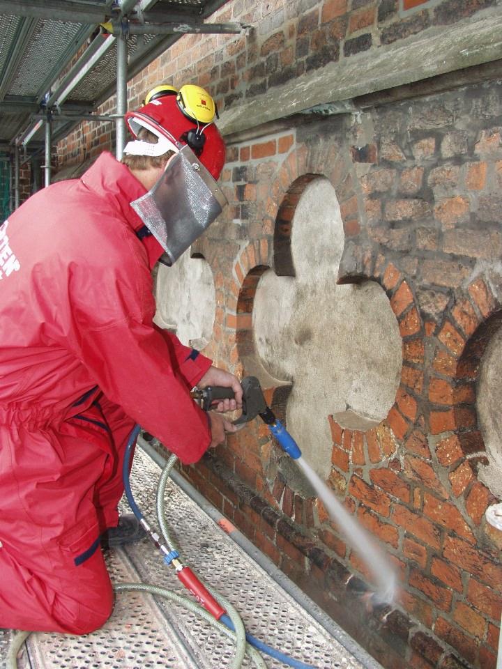 Pracownik w czerwonym kombinezonie i ochronnym hełmie hydropiaskuje ceglaną ścianę zabytkowego budynku z dekoracyjnymi elementami, stojąc na metalowym rusztowaniu.