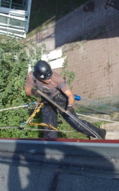 Pracownik w kasku i uprzęży alpinistycznej czyści rynnę na wysokości, używając łopatki i worka na odpady. Widoczny fragment dachu i elewacji budynku.