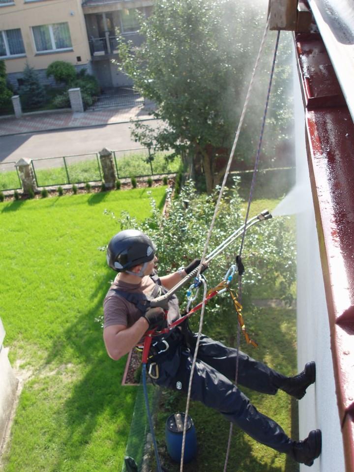 Pracownik w uprzęży alpinistycznej czyści elewację budynku strumieniem wody pod ciśnieniem, z widocznym efektem czyszczenia.