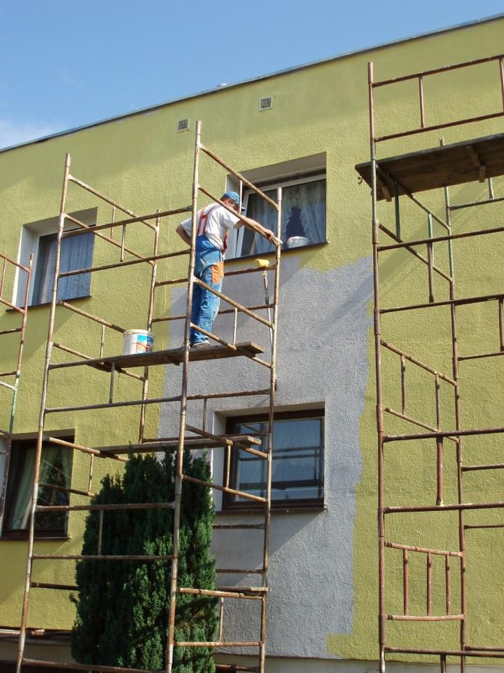 Mężczyzna w kombinezonie maluje elewację budynku na żółto, stojąc na rusztowaniu. Widoczny fragment pomalowanej na szaro ściany, kubełek z farbą i zielony krzew u podstawy rusztowania.