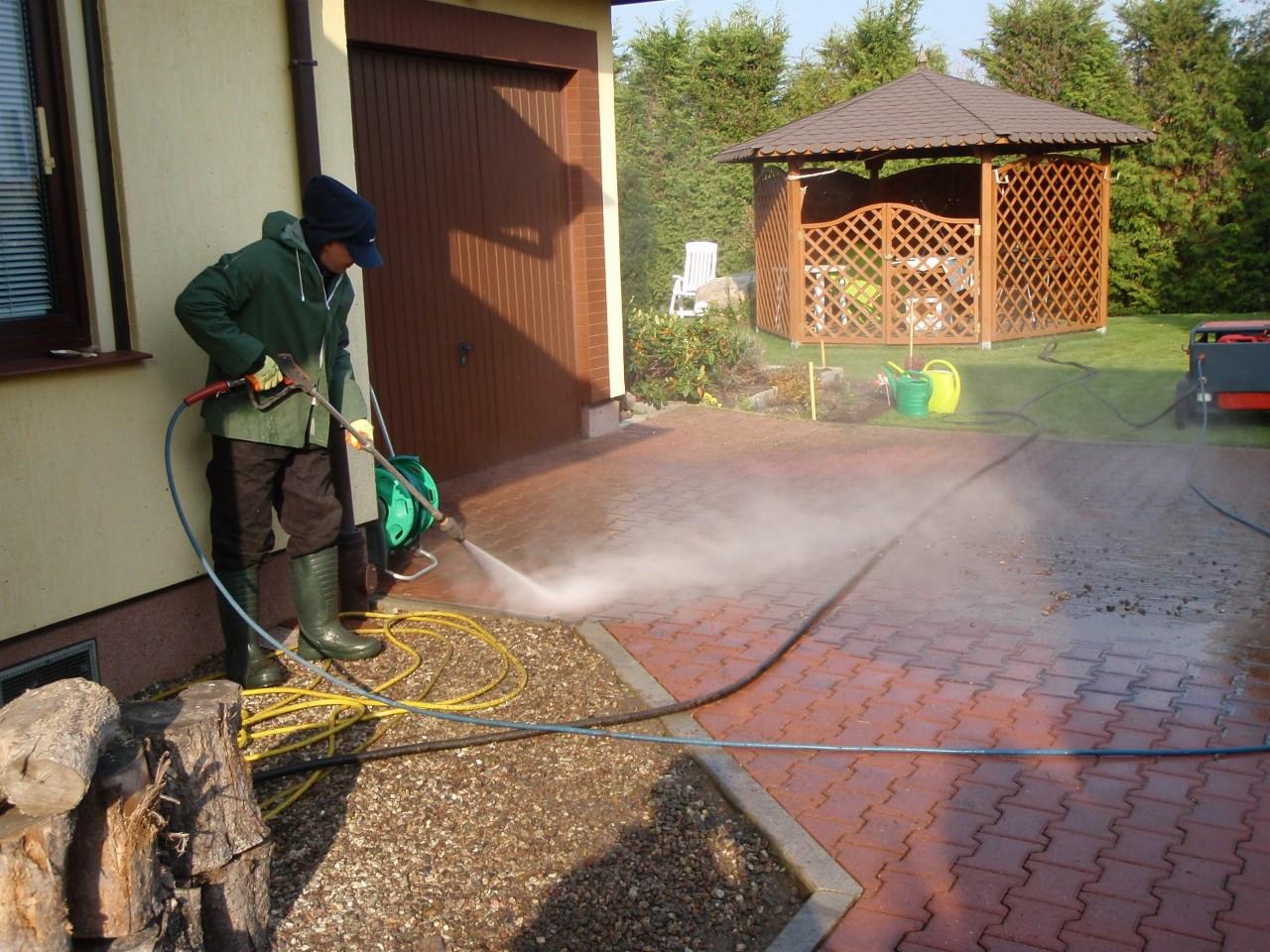 Mężczyzna w zielonej kurtce i kaloszach czyści czerwoną kostkę brukową na podjeździe myjką ciśnieniową; w tle widoczna altana ogrodowa i ściana budynku.