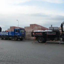 Dwa samochody ciężarowe z dźwigami, załadowane materiałami budowlanymi, stoją na placu wyłożonym kostką brukową. Widoczne palety z cegłami i innymi materiałami.
