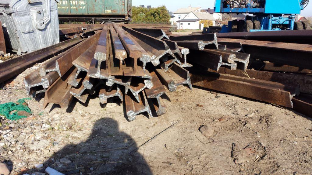 Stos zardzewiałych szyn kolejowych S49 na ziemi, w tle kontener, budynki i niebieski sprzęt budowlany. Widoczny cień osoby robiącej zdjęcie.