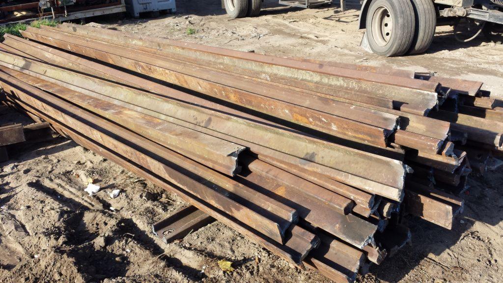 Stos szyn kolejowych S49, częściowo skorodowanych, ułożonych na ziemi obok pojazdu z widocznymi oponami.