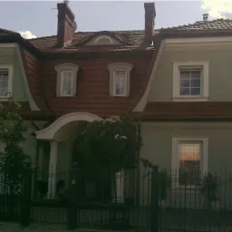 Zakończona budowa domu jednorodzinnego z dwuspadowym dachem pokrytym czerwoną dachówką, zieloną elewacją i białymi ramami okiennymi, widziana zza czarnego, metalowego ogrodzenia.