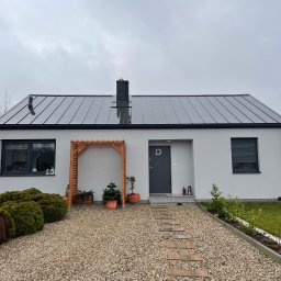 Alvic Investment - Parterowy dom z szarą, panelową blachodachówką, kremową elewacją, czarnymi ramami okiennymi i drewnianą pergolą nad wejściem, otoczony żwirowym podjazdem i zielenią.