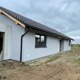 Nowoczesny dom jednorodzinny z szarą elewacją i czarną dachówką, widok z boku, fragment białego samochodu dostawczego w kadrze, teren wokół domu w trakcie prac ziemnych.