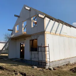 Surowy stan domu modułowego w trakcie budowy, widoczne ściany z płyt warstwowych, drewniane belki stropowe, drabina oparta o elewację, w tle budynek mieszkalny.