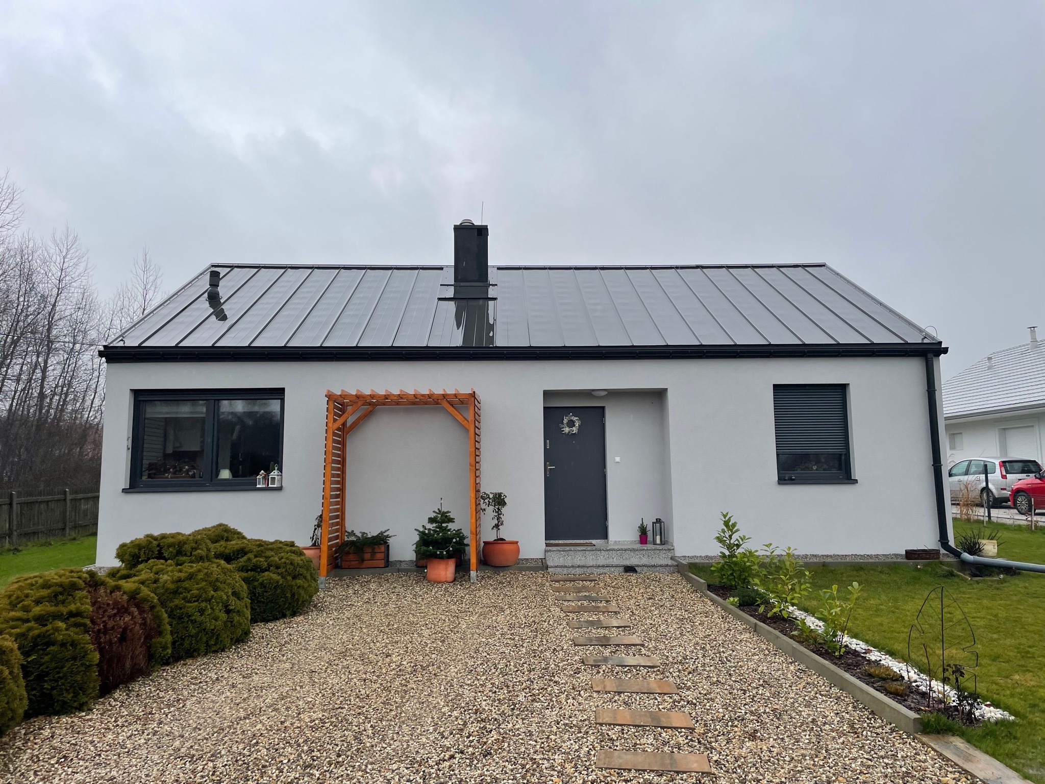 Parterowy dom z szarą, panelową blachodachówką, kremową elewacją, czarnymi ramami okiennymi i drewnianą pergolą nad wejściem, otoczony żwirowym podjazdem i zielenią.