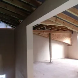 Wnętrze domu w trakcie prac tynkarskich: ściany pokryte świeżym, jasnym tynkiem, surowy drewniany strop z widoczną konstrukcją, fragment poddasza z płyt OSB, betonowa wylewka podłogowa.