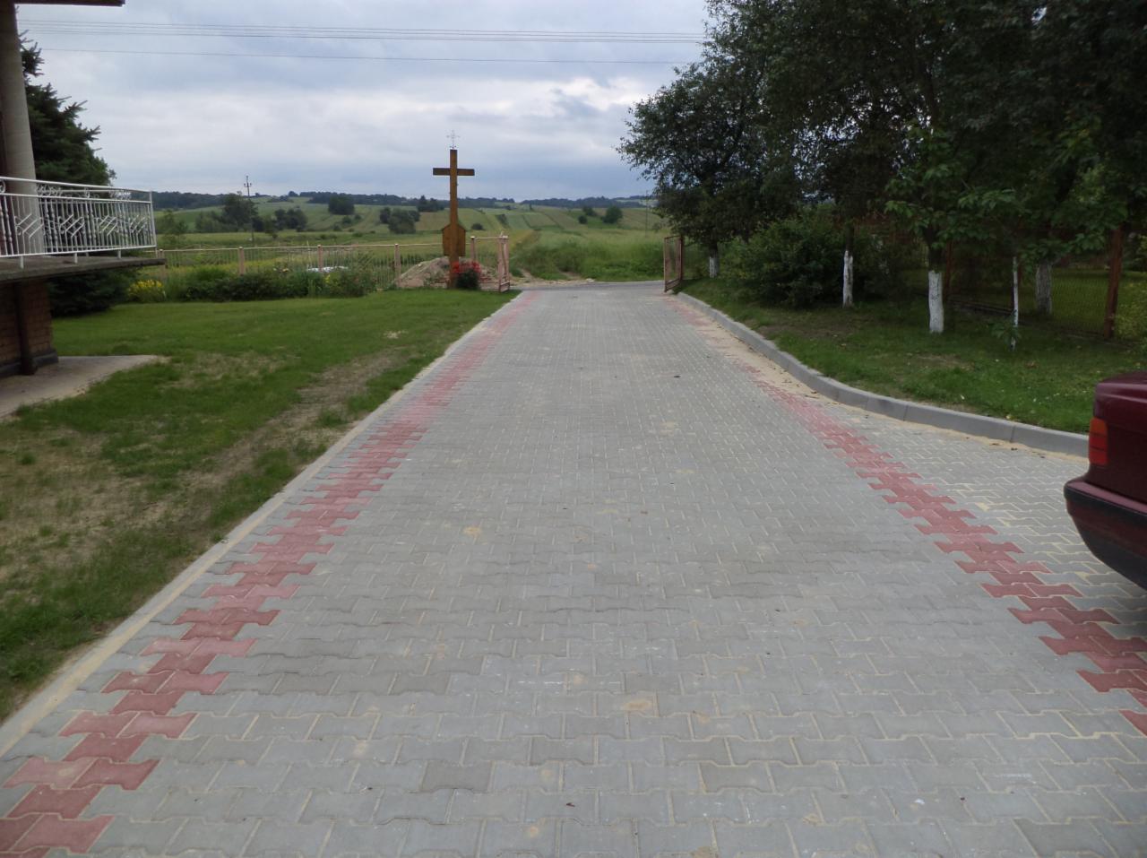 Szeroki podjazd z kostki brukowej w odcieniach szarości i czerwieni, prowadzący do drewnianego krzyża na tle zielonego krajobrazu. Widoczny fragment samochodu.