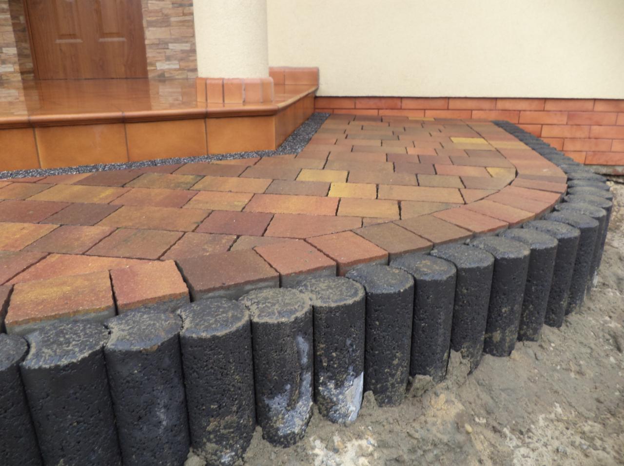 Kaskadowe ułożenie pomarańczowo-żółtej kostki brukowej, obrzeża z betonowych palisad, prowadzące do wejścia domu z kolumną i schodami.