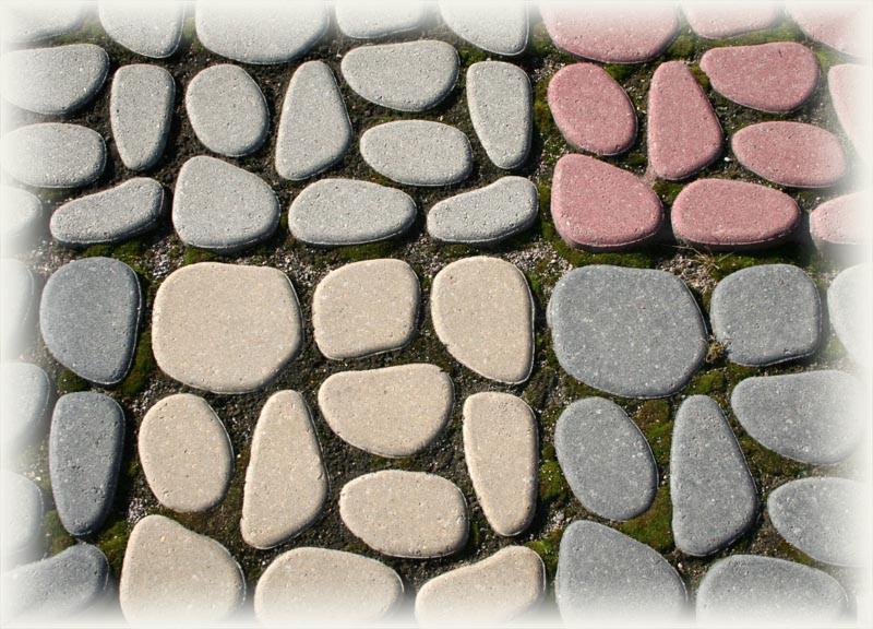 Nietypowa nawierzchnia z nieregularnych, zaokrąglonych elementów betonowych w odcieniach szarości, beżu i różu, z drobnymi porostami mchu w spoinach.