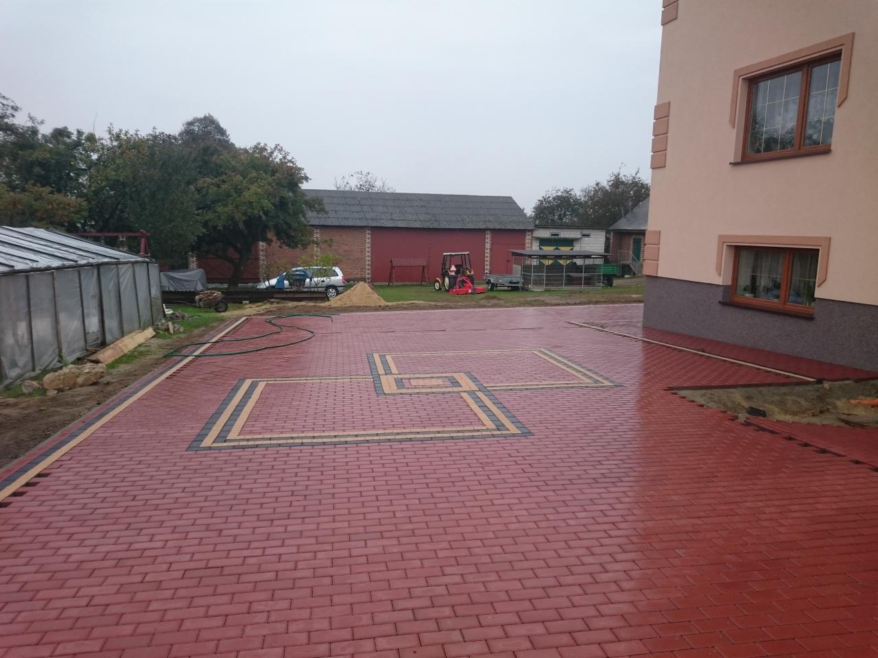 Czerwona kostka brukowa ułożona w geometryczny wzór na podwórku, obok domu z beżową elewacją i brązowymi oknami. W tle zabudowania gospodarcze i zieleń.