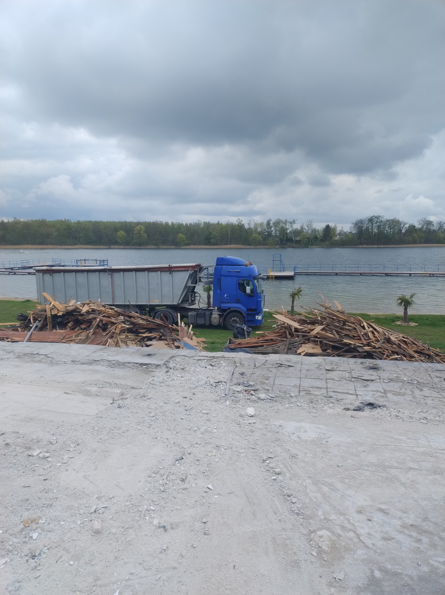 Ciężarówka z kontenerem obok stosu drewnianych odpadów budowlanych na tle jeziora i pochmurnego nieba.