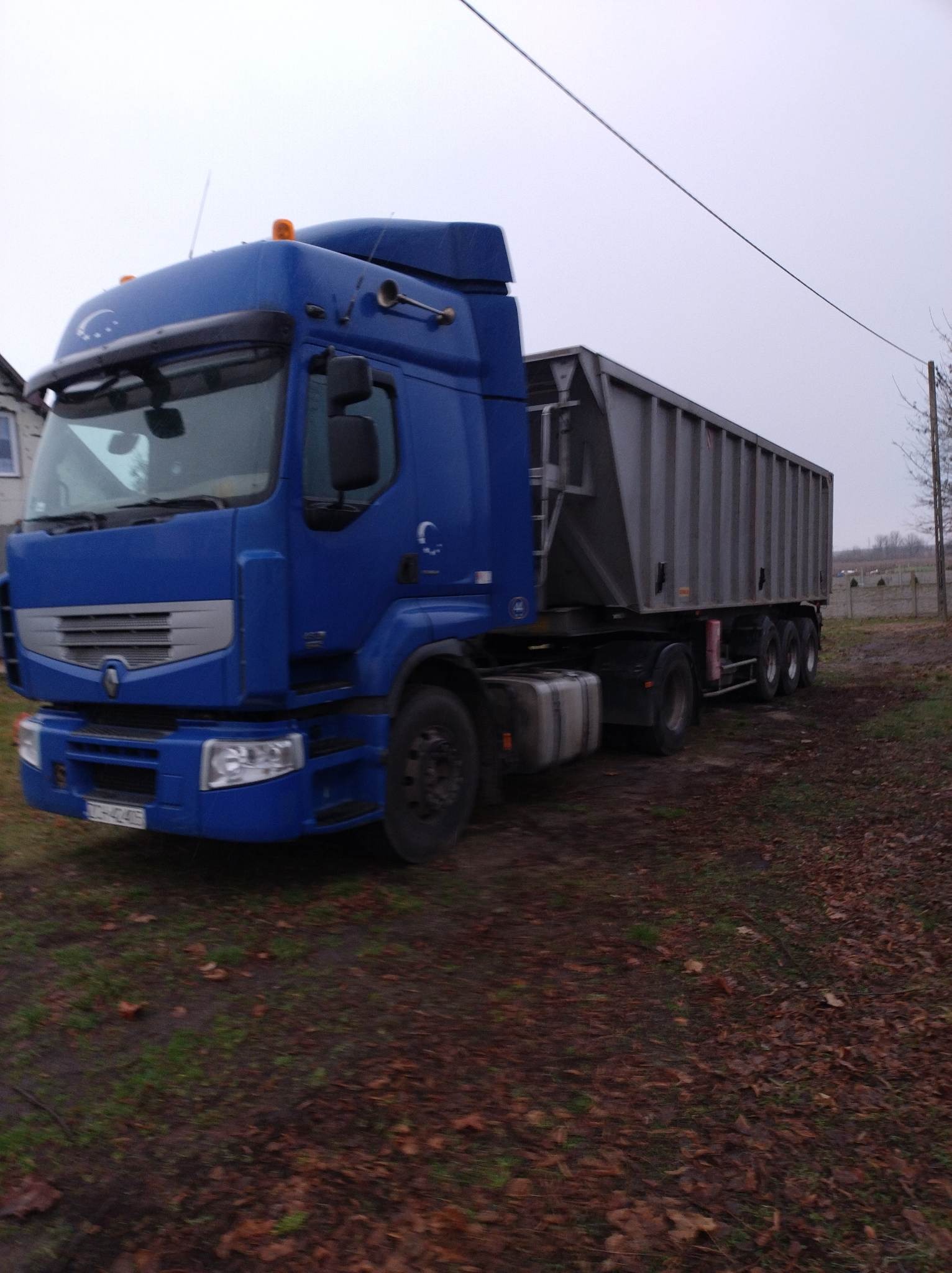 Niebieska ciężarówka Renault z przyczepą typu wanna, zaparkowana na ziemi pokrytej liśćmi, widoczna tablica rejestracyjna i logo firmy na kabinie.
