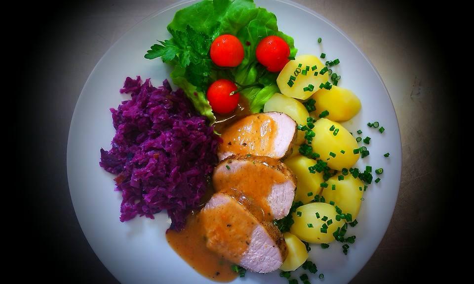 Elegancko podane danie na białym talerzu: plasterki mięsa w sosie, gotowane ziemniaki posypane szczypiorkiem, czerwona kapusta i sałatka z pomidorkami cherry.