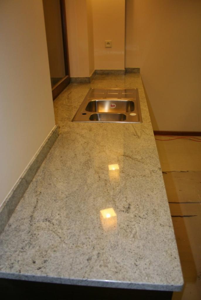 Jasny, granitowy blat kuchenny z wbudowanym, dwukomorowym zlewem ze stali nierdzewnej, widoczne odbicia światła na polerowanej powierzchni kamienia.