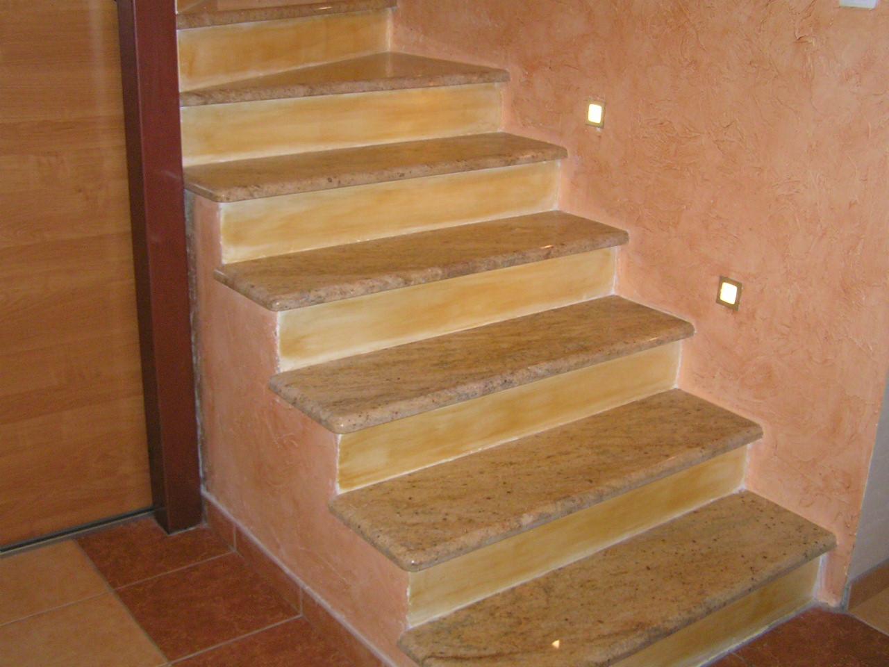 Granitowe schody wewnętrzne z oświetleniem punktowym w ścianie o fakturze trawertynu, widok z dołu.