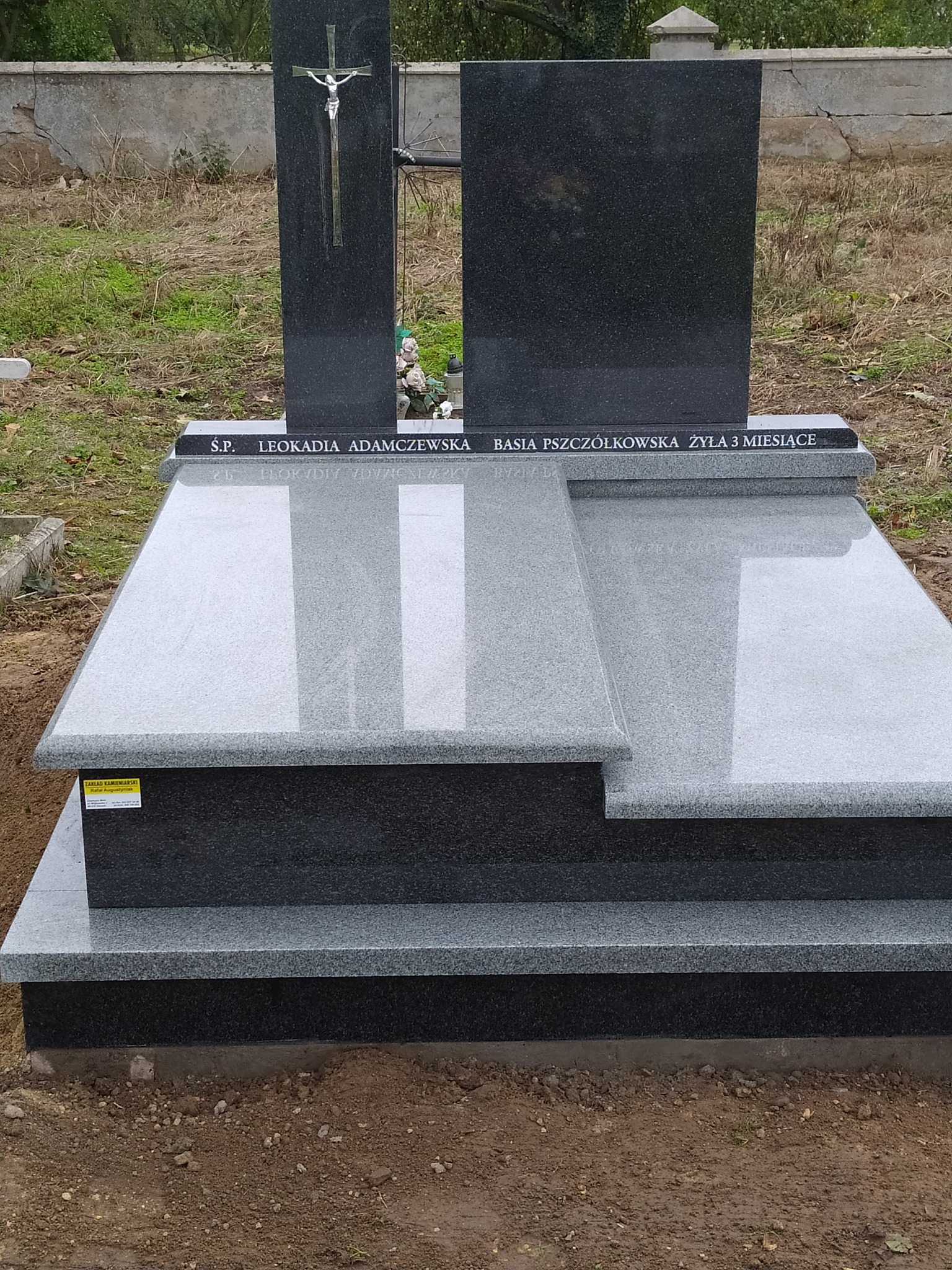 Granitowy nagrobek z krzyżem i inskrypcjami, widok z przodu, na cmentarzu.