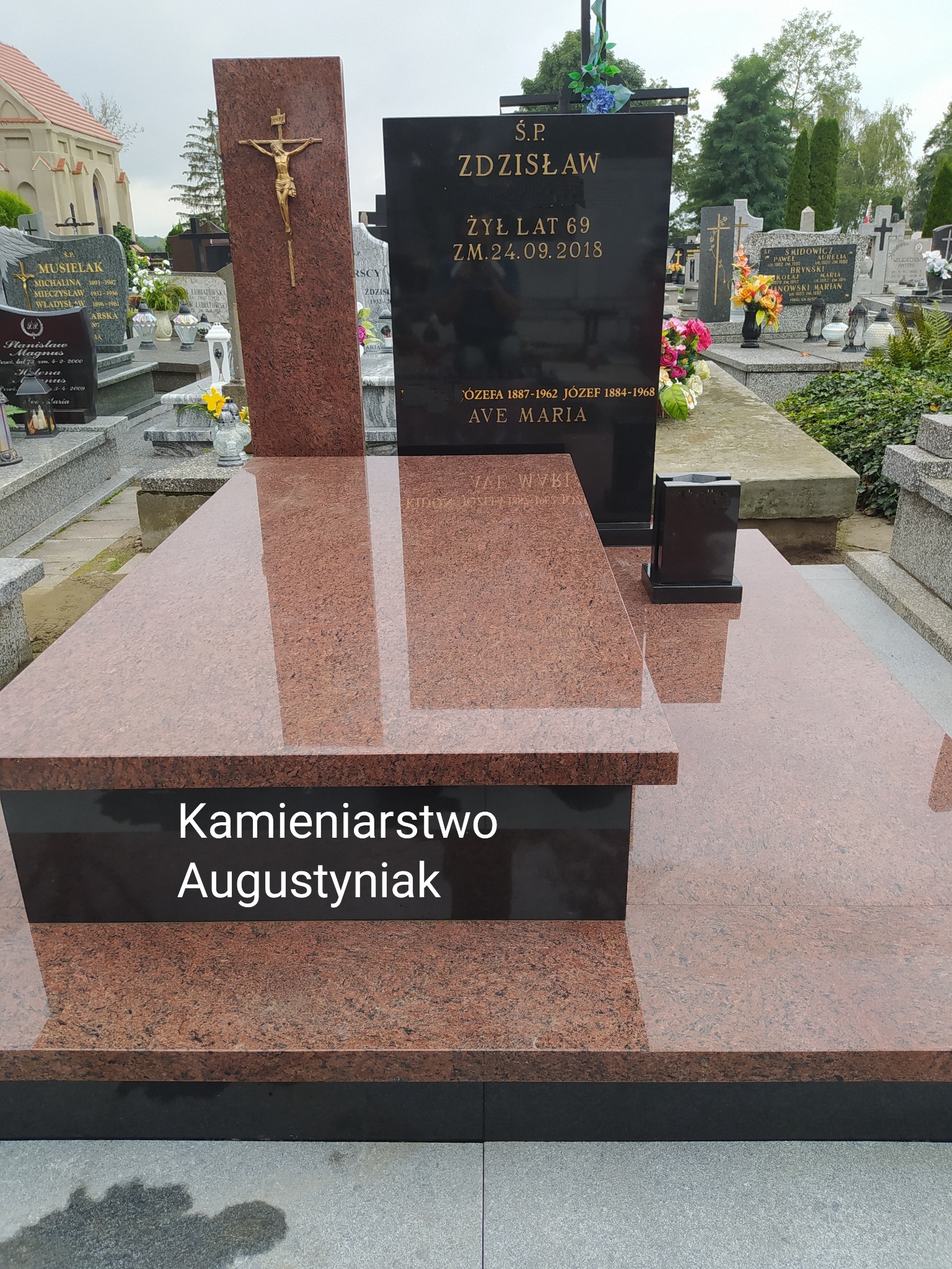 Nowoczesny nagrobek z czerwonego granitu z czarną tablicą nagrobną i złotym krucyfiksem, na tle innych grobów na cmentarzu.