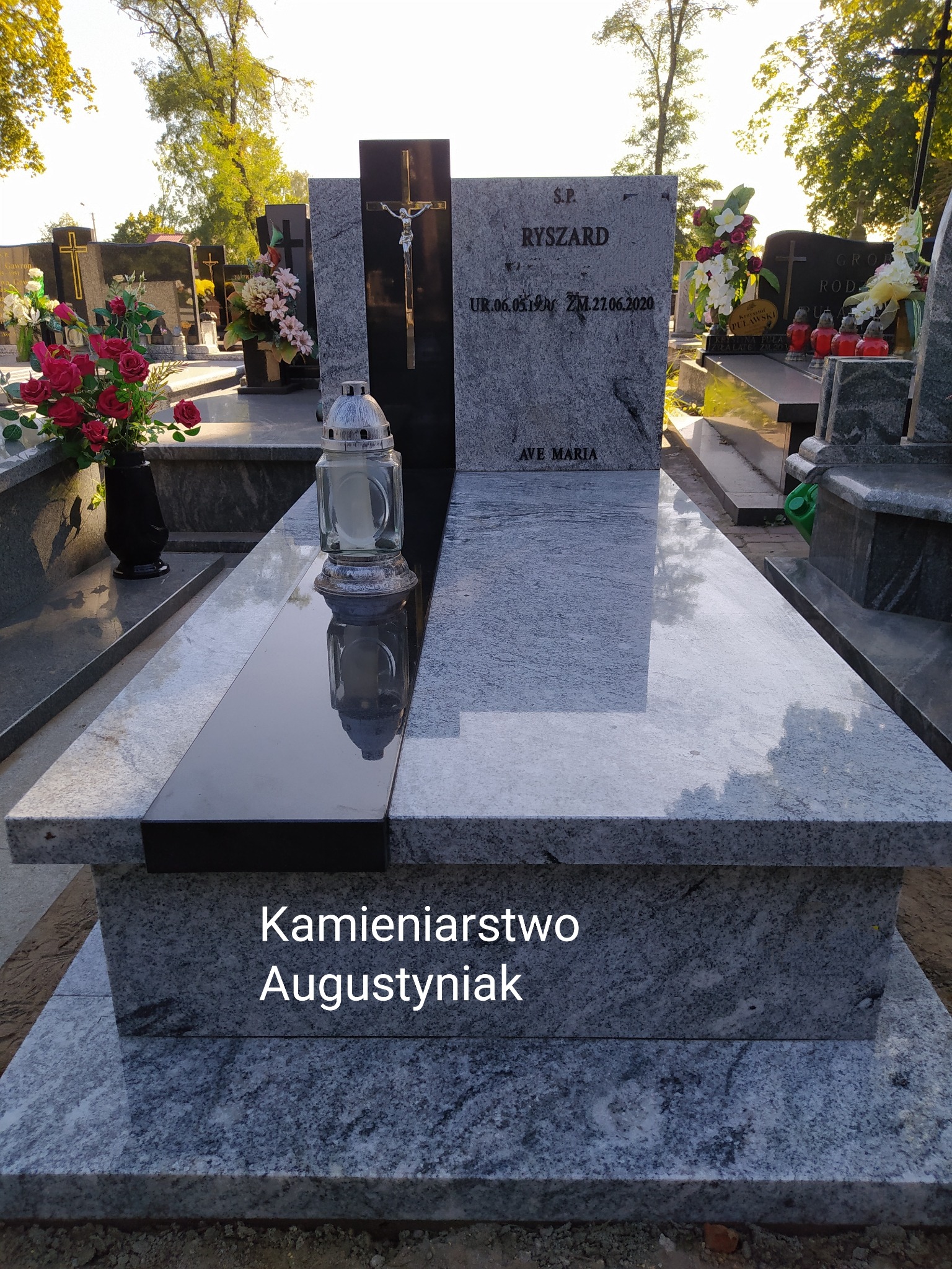 Granitowy nagrobek z czarnym akcentem, krzyżem i zniczem, w otoczeniu innych grobów na cmentarzu w słoneczny dzień.